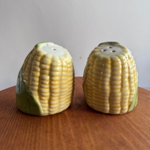 May include: A set of two ceramic salt and pepper shakers shaped like ears of corn. The shakers are yellow and white with green leaves.