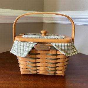 May include: A Longaberger Dresden Basket with a green and white plaid liner and a wooden lid. The basket has a brass tag that reads "Longaberger Dresden Basket 2001".