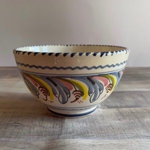 May include: A white ceramic bowl with a blue, yellow, green, and pink floral design. The bowl has a scalloped rim and a slightly flared base.
