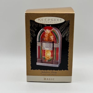 May include: A gold-toned box featuring a decorative ornament. The ornament is a miniature jukebox with red, green, and silver accents. The text "Keepsake Ornament" and "1996" are visible. The box also has the text "Jukebox Party Light and Music MAGIC".