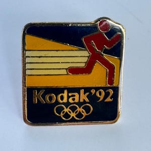 May include: A vintage Kodak '92 Olympic pin. The pin features a running figure in red, set against a yellow and blue background. The pin has the Olympic rings and the Kodak logo.