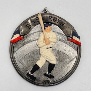 May include: A circular ornament featuring a baseball player in a vintage uniform, holding a bat. The ornament has a banner that reads "BABE RUTH" and includes red, white, and blue accents. The number "94" is at the bottom.