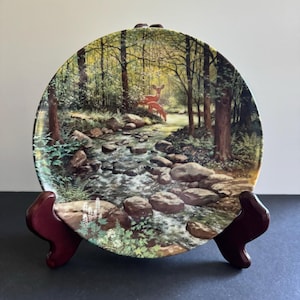 May include: A ceramic plate with a woodland scene featuring a stream flowing through a forest with rocks and two deer. The plate is on a wooden stand and has the word "JERNER" printed on the bottom.