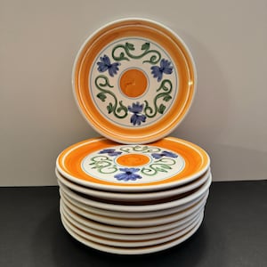 May include: A stack of ten white ceramic plates with a hand-painted orange, green, and blue floral design. The plates have a white rim and a slightly raised edge.