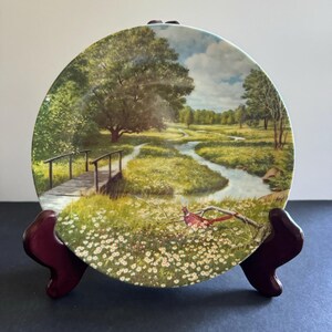 May include: A decorative ceramic plate with a painting of a winding stream, a wooden bridge, a green meadow, and a pheasant in a field of white flowers. The plate is on a brown stand.