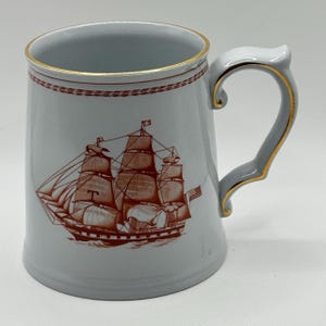 May include: A light gray ceramic mug with a brown sailing ship illustration. The mug has a gold rim and handle accents. The handle is gray with gold trim. The ship illustration includes the American flag.