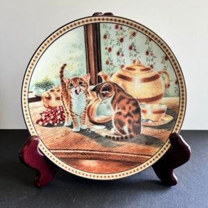May include: A decorative ceramic plate with a brown border and a white center featuring a painting of two kittens. One kitten is brown and white and is looking down at a spilled bowl of milk. The other kitten is orange and white and is looking at the viewer with a surprised expression. The plate is on a wooden stand.