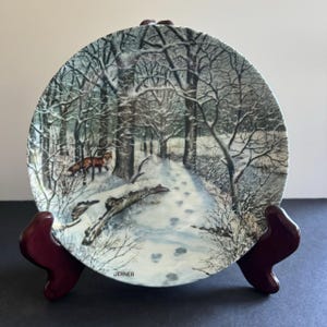 May include: A white ceramic plate with a winter scene of a snowy forest path with footprints. The plate features a brown fox walking through the snow. The plate is signed "JERNER" in the bottom center.