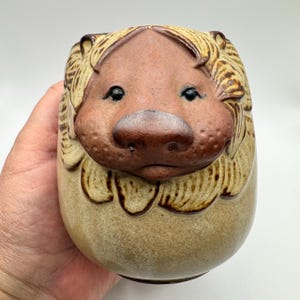 May include: A ceramic lion-shaped mug with a tan base and a detailed face. The lion's face is brown with black eyes and a prominent nose. The mane is a lighter tan color, and the mug has a rounded, egg-like shape.