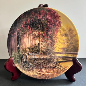 May include: A decorative ceramic plate with a painting of a rustic scene. The plate features a wooden fence, a weathered barn, a rusty wheelbarrow, and a lush green vine with red flowers. The background is a soft yellow with a hint of orange.