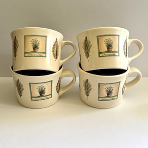 May include: Four cream-colored ceramic mugs stacked together. Each mug features a square image of a potted plant with the word "chamomile" below it. A leaf design is on the side of each mug. The mugs have a black interior and a curved handle.