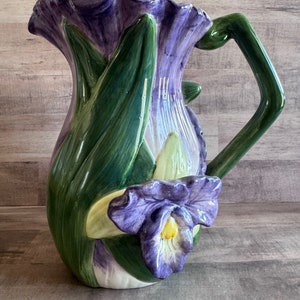 May include: A ceramic pitcher with a purple and green floral design. The pitcher has a wide mouth and a handle that is shaped like a leaf. The pitcher is decorated with a large purple flower with a yellow center.