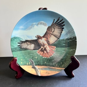 May include: A decorative ceramic plate featuring a red-tailed hawk in flight against a backdrop of a mountain range and a forest. The plate is on a wooden stand.