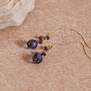 May include: A pair of dangle earrings featuring blue glass beads and purple flower accents. The earrings are suspended from a delicate gold chain.