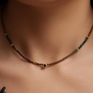 May include: A delicate necklace featuring a brown beaded chain with small green and gold accents. A single black stone is centered on the necklace. The necklace is worn around the neck.