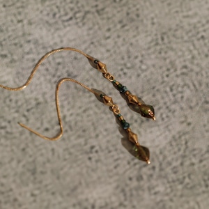 May include: A pair of gold threader earrings. Each earring has a fine chain and a cluster of small, faceted beads in shades of green and gold. The earrings are shown on a textured, grey surface.