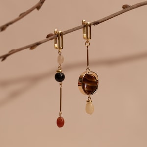May include: Two gold-colored earrings with dangling elements. One earring features a series of beads in black, pink, and red. The other earring has a circular pendant with a brown stone and a teardrop-shaped bead.