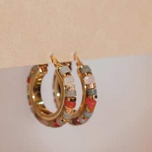Puede incluir: Un par de pendientes de aro dorados con cuentas geométricas y coloridas. Las cuentas son en tonos rojos, naranjas, grises y rosas. Los pendientes cuelgan sobre un fondo marrón claro.