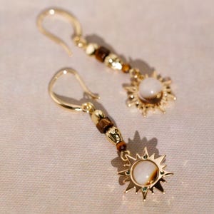 Op de afbeelding: Gouden oorbellen met een zonnestralenontwerp en een witte steen in het midden. De oorbellen zijn versierd met bruine kralen.