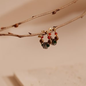 May include: A pair of hoop earrings featuring a gold-tone metal wire adorned with small wooden beads, smooth red spherical beads, a single green bead, and a larger central bead with a striped pattern in shades of gray, black, and white.