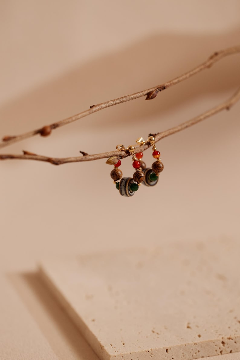 May include: A pair of hoop earrings featuring a gold-tone metal wire adorned with small wooden beads, smooth red spherical beads, a single green bead, and a larger central bead with a striped pattern in shades of gray, black, and white.