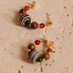 May include: A pair of hoop earrings featuring gold-tone metal wire strung with a variety of beads, including small red spheres, dark brown textured wooden spheres, a small green bead, and a larger central bead with blue, white, and gray stripes.