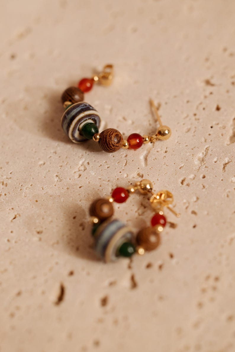 May include: A pair of hoop earrings featuring gold-tone metal wire strung with a variety of beads, including small red spheres, dark brown textured wooden spheres, a small green bead, and a larger central bead with blue, white, and gray stripes.