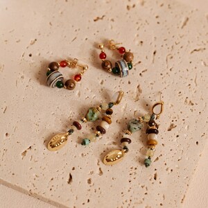 May include: Two pairs of earrings featuring gold-tone metal hardware. One pair consists of small hoops adorned with multicolored wooden, ceramic, and stone beads. The second pair features dangling strands of assorted natural stones and gold-tone charms.