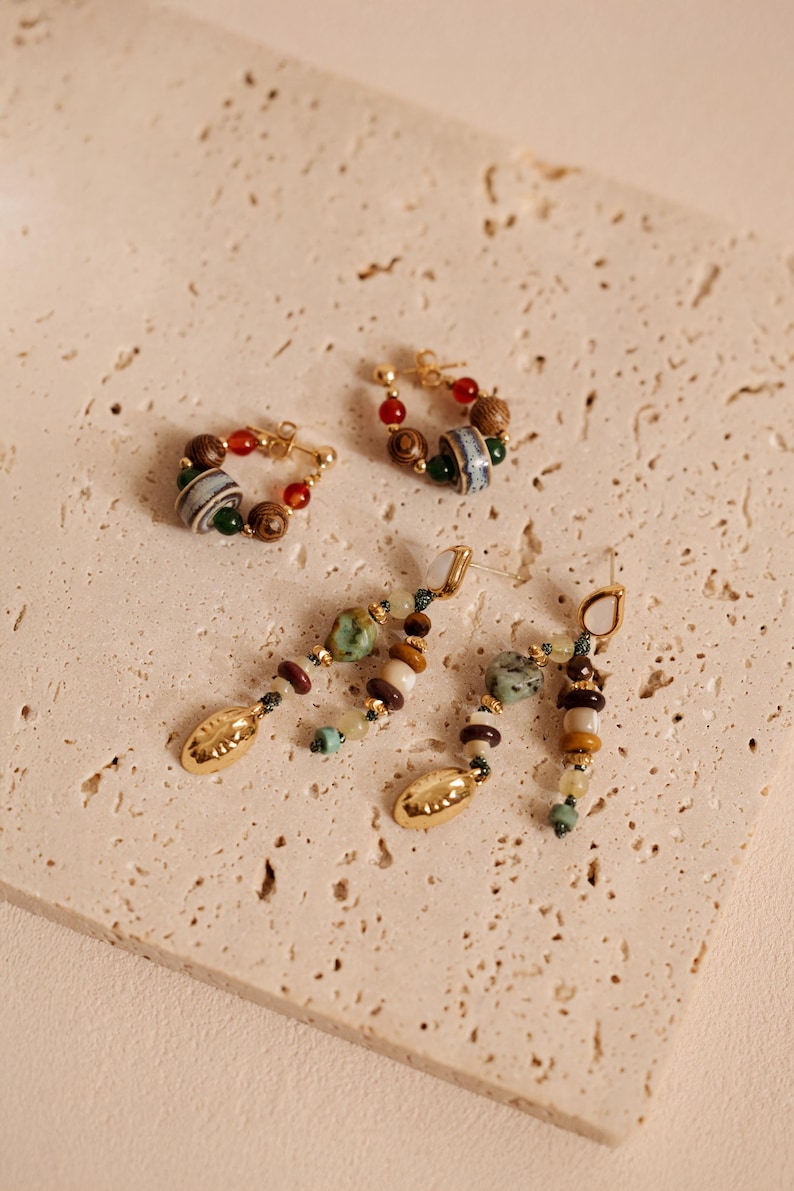 May include: Two pairs of earrings featuring gold-tone metal hardware. One pair consists of small hoops adorned with multicolored wooden, ceramic, and stone beads. The second pair features dangling strands of assorted natural stones and gold-tone charms.
