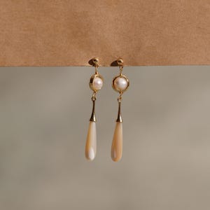 May include: A pair of elegant gold-toned earrings. Each earring features a round pearl set in a gold frame, with a long, teardrop-shaped, cream-colored pendant. The earrings are displayed against a neutral background.