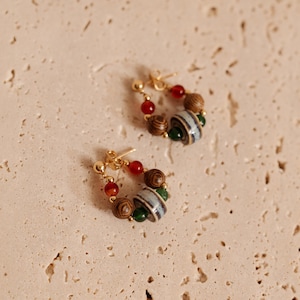 May include: A pair of hoop earrings featuring gold-tone metal posts and a collection of beads. Each earring includes a central grey and white striped ceramic bead, flanked by dark brown wood-grain beads, small green beads, and translucent red beads.