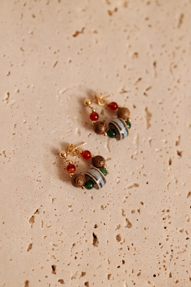May include: A pair of hoop earrings featuring gold-tone metal posts and a collection of beads. Each earring includes a central grey and white striped ceramic bead, flanked by dark brown wood-grain beads, small green beads, and translucent red beads.