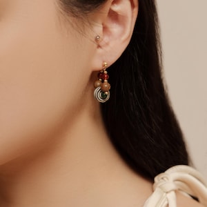 May include: A drop earring featuring a small gold-tone stud, a deep red bead, a light brown wooden bead with a dark grain pattern, and a circular charm with concentric white, gray, and green rings hanging from a gold-tone metal chain link.