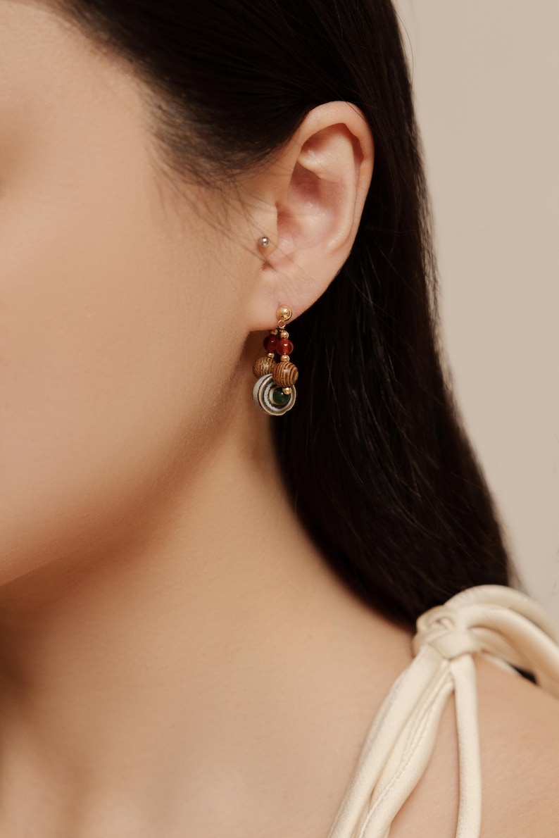 May include: A drop earring featuring a small gold-tone stud, a deep red bead, a light brown wooden bead with a dark grain pattern, and a circular charm with concentric white, gray, and green rings hanging from a gold-tone metal chain link.