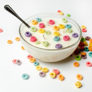 May include: A white bowl filled with a white candle shaped like a bowl of cereal. The candle is decorated with colorful cereal rings. A silver spoon is resting on the rim of the bowl. Scattered around the bowl are more colorful cereal rings.