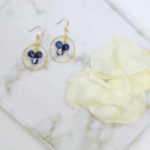 Puede incluir: Un par de pendientes de aro dorados, cada uno con tres cuentas moteadas azules y blancas. Los pendientes se exhiben en una bandeja de m&aacute;rmol blanco con p&eacute;talos de flores blancas.