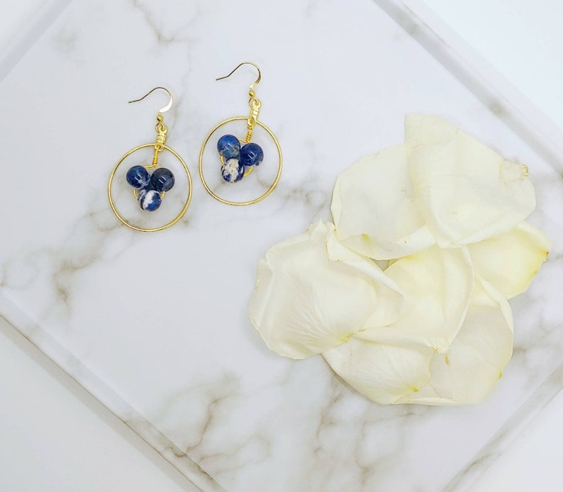 Puede incluir: Un par de pendientes de aro dorados, cada uno con tres cuentas moteadas azules y blancas. Los pendientes se exhiben en una bandeja de m&aacute;rmol blanco con p&eacute;talos de flores blancas.