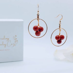 Puede incluir: Un par de pendientes de aro dorados con tres cuentas rojas agrupadas. Los pendientes se exhiben con una caja blanca que tiene el texto "Healing Jewelry Co." y un dise&ntilde;o floral. El fondo es blanco.