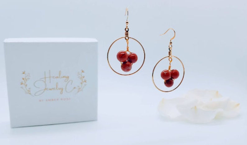 Puede incluir: Un par de pendientes de aro dorados con tres cuentas rojas agrupadas. Los pendientes se exhiben con una caja blanca que tiene el texto "Healing Jewelry Co." y un dise&ntilde;o floral. El fondo es blanco.