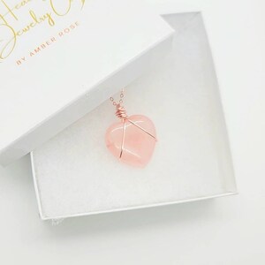 May include: A rose quartz heart pendant necklace in a white gift box. The heart-shaped stone is wrapped in copper wire and hangs from a delicate chain. The box has the text "Healing Jewelry Co. by Amber Rose" in gold lettering.