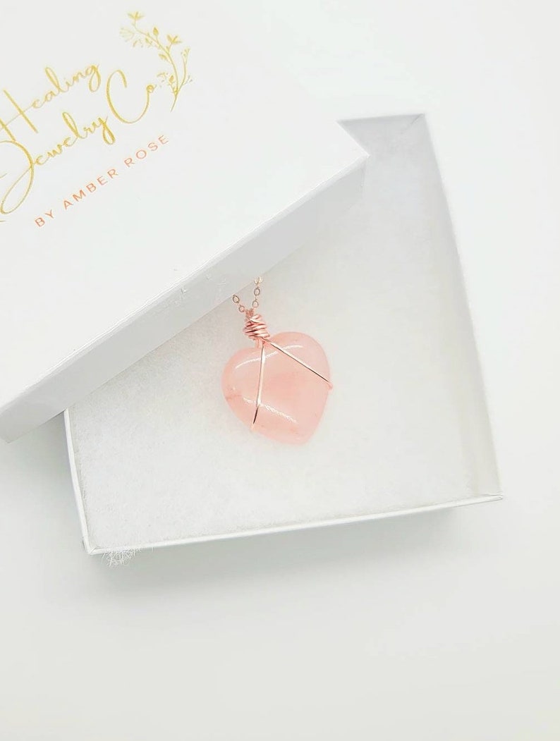 May include: A rose quartz heart pendant necklace in a white gift box. The heart-shaped stone is wrapped in copper wire and hangs from a delicate chain. The box has the text "Healing Jewelry Co. by Amber Rose" in gold lettering.