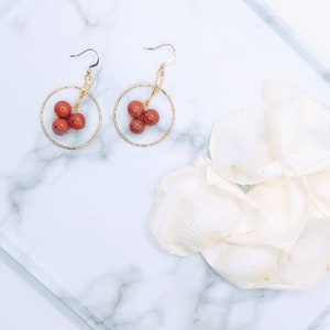 Puede incluir: Un par de pendientes de aro dorados, cada uno con un grupo de cuentas de color marr&oacute;n rojizo. Los pendientes se exhiben sobre una superficie de m&aacute;rmol blanco, con p&eacute;talos de flores blancas en el fondo. Dise&ntilde;o sencillo y elegante.