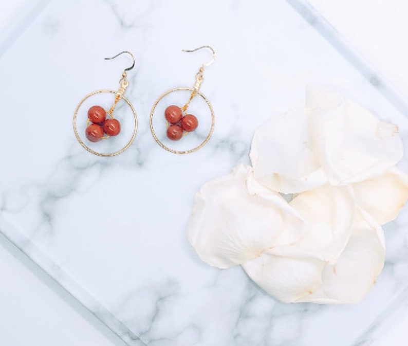 Puede incluir: Un par de pendientes de aro dorados, cada uno con un grupo de cuentas de color marr&oacute;n rojizo. Los pendientes se exhiben sobre una superficie de m&aacute;rmol blanco, con p&eacute;talos de flores blancas en el fondo. Dise&ntilde;o sencillo y elegante.