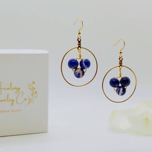 Puede incluir: Pendientes de aro dorados con tres cuentas moteadas en azul oscuro y blanco. Los pendientes se muestran junto a una caja blanca con el texto dorado "Healing Jewelry Co. by Amber Rose."
