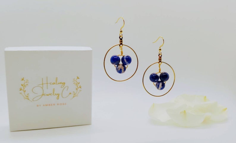 Puede incluir: Pendientes de aro dorados con tres cuentas moteadas en azul oscuro y blanco. Los pendientes se muestran junto a una caja blanca con el texto dorado "Healing Jewelry Co. by Amber Rose."