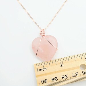 May include: A rose gold necklace with a heart-shaped rose quartz pendant. The pendant is approximately 1 inch wide and is wrapped in rose gold wire. The necklace chain is also rose gold and is displayed on a white background.
