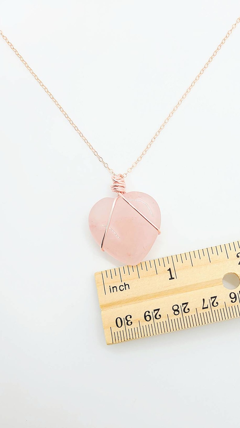 May include: A rose gold necklace with a heart-shaped rose quartz pendant. The pendant is approximately 1 inch wide and is wrapped in rose gold wire. The necklace chain is also rose gold and is displayed on a white background.