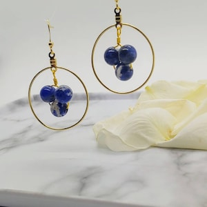 Puede incluir: Pendientes de aro dorados con tres cuentas moteadas azules y blancas suspendidas dentro de cada aro. Los pendientes tienen un dise&ntilde;o simple y elegante, perfectos para el uso diario o para ocasiones especiales. Se exhiben sobre una superficie de m&aacute;rmol blanco.