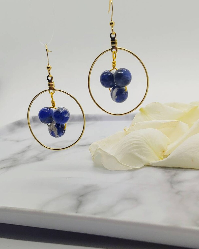 Puede incluir: Pendientes de aro dorados con tres cuentas moteadas azules y blancas suspendidas dentro de cada aro. Los pendientes tienen un dise&ntilde;o simple y elegante, perfectos para el uso diario o para ocasiones especiales. Se exhiben sobre una superficie de m&aacute;rmol blanco.