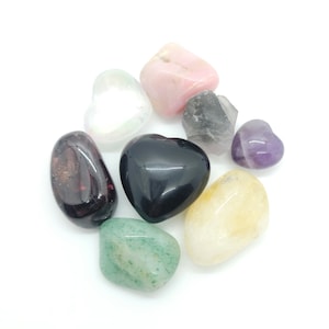 May include: An assortment of polished gemstones in various shapes and colors. Includes a black heart-shaped stone, a pink stone, a green stone, and a purple heart-shaped stone. The stones are arranged on a white background.